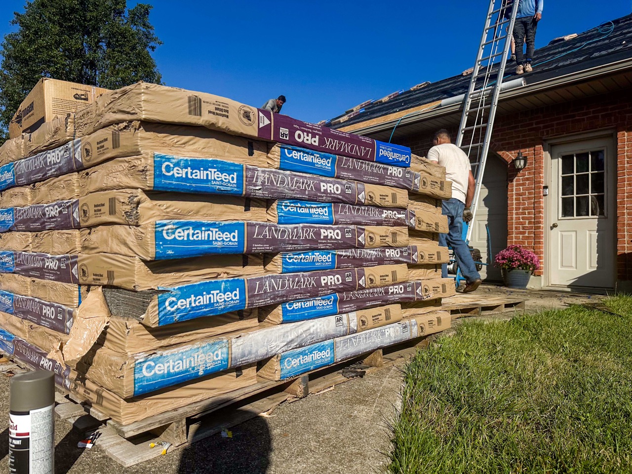 a pallet of Certainteed Landmark Shingles is focused in the foreground while roof technicians and a single story brick home is in the background