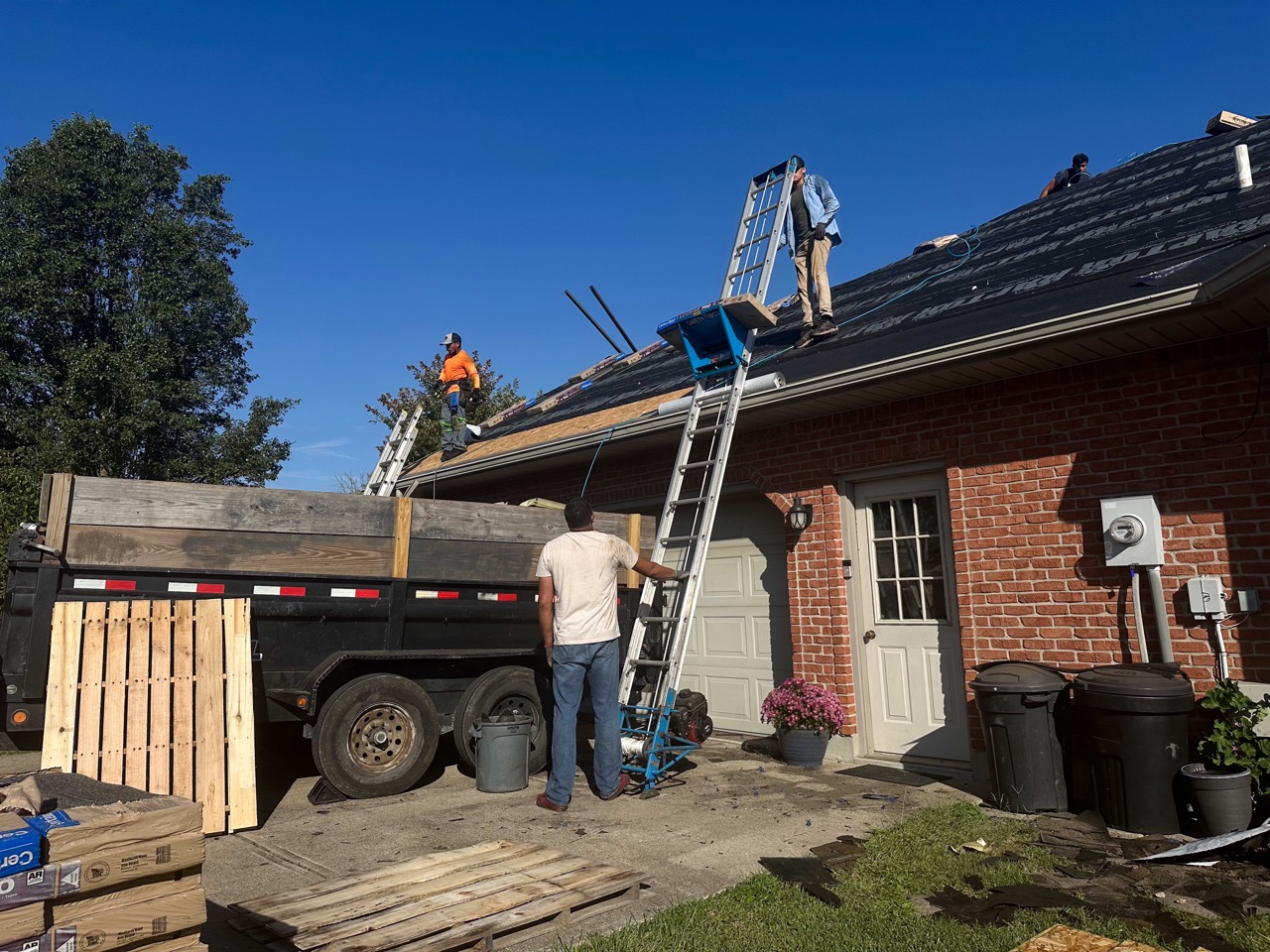 a ladder is setup on the side of a single story brick house while roof technicians are working on the roof and gathering supplies from the ground.