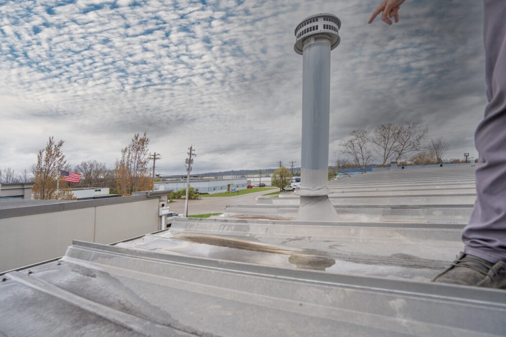 on top of gray standing seam metal roof on an industrial building. edge of frame shows the leg of an inspector and arm pointing out a roof vent needing possible attention